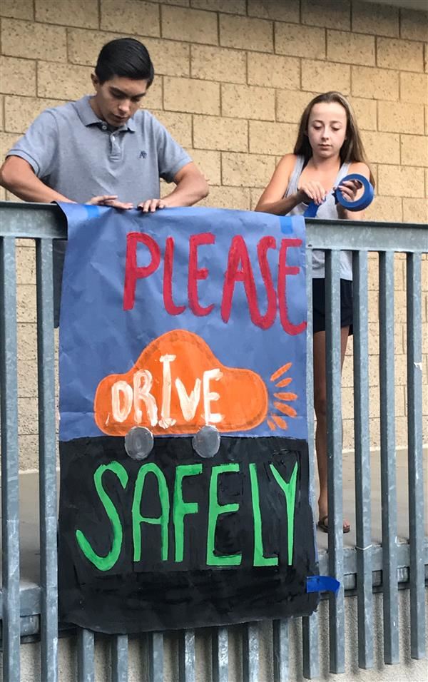 A high school boy and girl post a banner from a second story railing. The sign says Please Drive Safely A high school boy and girl post a banner from a second story railing. The sign says Please Drive Safely